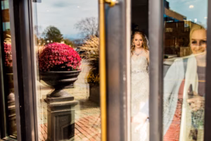 A bride in a white dress stands indoors near a glass door, with her reflection and outdoor plants visible through the glass.