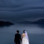 A bride and groom in formal attire stand holding hands by a large, calm lake under a cloudy, dramatic sky, facing away from the camera.