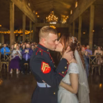 A bride and groom share a kiss indoors at their wedding ceremony, with guests seated in the background watching.