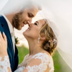A bride and groom stand close together, smiling and touching noses, with a wedding veil draped around them outdoors.