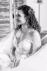 A bride with curly hair sits on a couch, wearing an off-the-shoulder lace wedding dress and a necklace.
