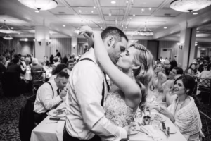 A bride and groom kiss while dancing closely at their wedding reception, surrounded by seated guests in a large, decorated banquet hall.