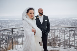 A woman in a white hooded cloak and a man in a suit stand on a snowy overlook with railings, with a wintry landscape in the background.
