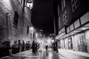 A bride and groom walk hand in hand down a wet alley at night, surrounded by guests holding sparklers, with brick buildings and shadows visible on both sides.