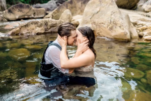 A man and a woman, both fully clothed, sit in a shallow rocky river, holding each other closely and touching foreheads.