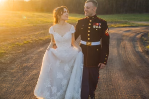 A bride in a white lace dress walks with a groom in a formal military uniform on a dirt road at sunset.