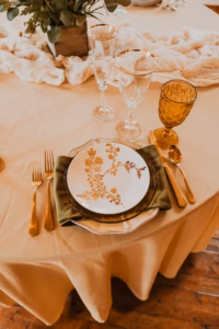 A formal table setting with gold flatware, a patterned plate on a green napkin, an amber goblet, two clear glasses, and a beige tablecloth with a centerpiece of greenery.