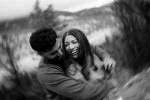 A man and woman embrace and laugh together outdoors, with a blurred natural background of trees and mountains. The image is in black and white.
