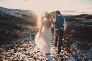 A bride and groom walk along a snowy path in a grassy landscape at sunset, with the sun shining between distant hills.