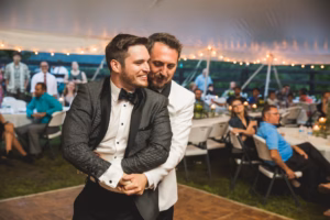 Two men in tuxedos dance closely together at an outdoor event under a tent, with seated guests and string lights in the background.