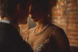A man and woman stand close to each other in low light, with the woman's face partially shadowed. She wears a pearl necklace and an ornate dress. A brick wall is in the background.