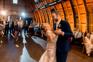 A bride and groom share their first dance in a wooden barn while wedding guests watch in the background.