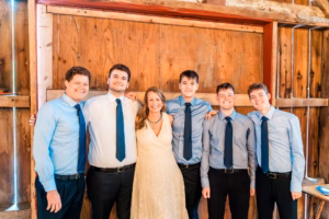 Six people pose indoors in front of a wooden wall; five men in blue shirts and ties stand around a woman in a cream dress.