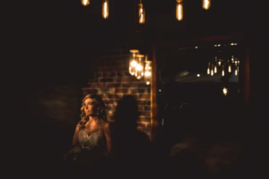 A woman in a wedding dress stands by a brick wall, illuminated by hanging light bulbs in a dimly lit room.