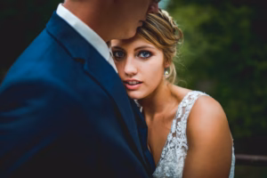 A woman in a white lace dress looks at the camera while embracing a man in a navy suit outdoors.