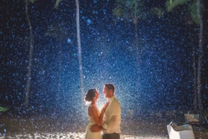 A couple stands on a sandy beach at night, illuminated by bright backlighting as raindrops or snow fall around them.