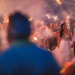 A bride and groom walk through a crowd of guests holding sparklers at night, with smoke and lights creating a festive atmosphere.