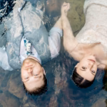 A man in a vest and tie and a woman in a lace dress lie side by side, holding hands, partially submerged in clear water with rocks visible below.