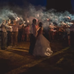 A bride and groom stand facing each other outdoors at night, surrounded by guests holding sparklers, with smoke and lights creating a festive atmosphere.
