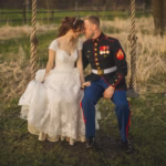 A bride in a white dress and a groom in a military uniform sit closely together on a wooden swing outdoors, touching foreheads.