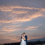 A couple in wedding attire embraces outdoors at sunset, with a dramatic cloudy sky and distant hills in the background.