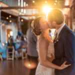 A bride and groom kiss on the dance floor at their wedding reception, with sunlight shining behind them and guests seated in the background.