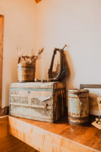 A rustic wooden chest, a metal container, a bucket of dried wheat, and an oval mirror sit on a wooden surface against a cream-colored wall.