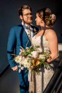 A bride in a lace dress holding a bouquet stands next to a groom in a blue suit and floral tie; they look at each other and smile in natural light.