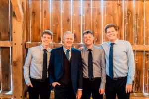 Four men in dress shirts and ties, one in a suit, stand together smiling in front of a wooden wall with string lights.