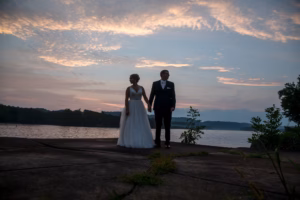 A bride and groom hold hands while walking by a lakeside at sunset, with trees and a cloudy sky in the background.