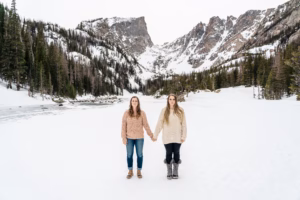 Two women stand holding hands on a snowy landscape with mountains and pine trees in the background.