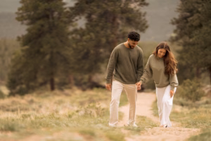 A man and woman walk hand-in-hand on a dirt path in a forested area, both wearing green sweaters and light pants.