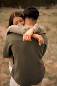 A woman hugs a man outdoors, displaying an engagement ring on her finger with her eyes closed.