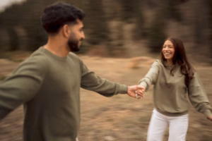 A man and woman wearing green sweaters hold hands and smile at each other outdoors, with a blurred forest background.