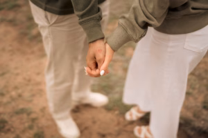 Two people holding hands, each with a small red heart drawn on their hands. They are standing outdoors, wearing light-colored pants and greenish sweaters.