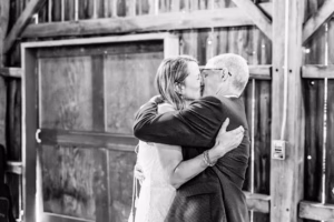 A bride and an older man embrace and kiss on the cheek in a rustic wooden venue, both wearing formal attire.
