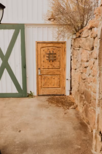 A wooden door with metal studs and a small barred window, set in a white wall next to a green-trimmed barn door and a stone wall.