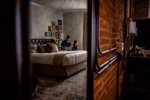 A person styles another person's hair in a dimly lit bedroom with a large bed and framed pictures on the wall.