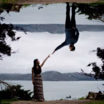 A woman and a man reach toward each other, with the man appearing upside down due to a mirror effect, near a lake with trees and mountains in the background.