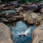 A man and woman in formal clothing float on their backs in a small pool of water surrounded by large rocks and a forested area.