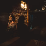 A bride in a white dress sits by a brick wall in dim lighting, illuminated by a soft spotlight with hanging light bulbs visible above.