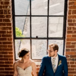 A bride and groom stand holding hands in front of a large window set in a brick wall, looking at each other.