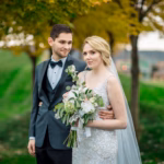 A bride in a white dress holds a bouquet while standing next to a groom in a tuxedo, outdoors with green grass and trees in the background.
