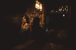 A bride in a white dress sits in dim lighting against a brick wall, holding a bouquet, with soft light illuminating her face and upper body.