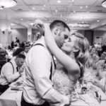A bride and groom share a kiss on the dance floor at their wedding reception, surrounded by seated guests in a banquet hall.