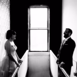 A bride and groom stand facing each other at opposite sides of a narrow hallway, with a large window behind them letting in natural light.