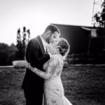 A bride and groom share a kiss outdoors at sunset, both dressed in formal wedding attire.