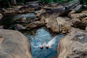 A man and a woman in formal attire float on their backs in a shallow rocky stream surrounded by large boulders and greenery.