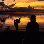 A silhouetted man skips a stone across a calm lake at sunset while a silhouetted woman stands watching in the foreground.