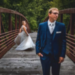 A bride stands on a wooden bridge in the background, while a groom in a blue suit stands in the foreground, slightly out of focus. Trees surround the bridge.